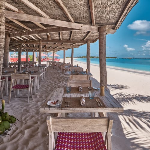 Outdoor dining area under a thatched roof on a sandy beach with wooden tables and chairs set with plates and glasses, overlooking a calm blue ocean and clear sky.