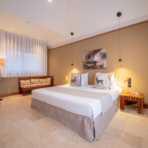 Ein modernes Hotelzimmer mit einem großen, ordentlich gemachten Bett mit weißen Bettlaken und zwei dekorativen Kissen mit Tiermotiven, warmem Licht von Nachttischlampen, einer Holzbank unter einem Fenster mit weißen Vorhängen und einer gemütlichen Sitzecke mit Stuhl und Lampe.