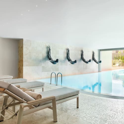Innenbereich eines Spa-Pools mit mehreren Liegestühlen neben einem rechteckigen Pool, der wandmontierte Tauchskulpturen zeigt, mit einem großen Fenster mit Blick auf die Außenbegrünung.