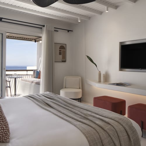Modern hotel room with a large bed, patterned pillows, gray blanket, flat-screen TV mounted on the wall, and a balcony overlooking the sea with outdoor seating.