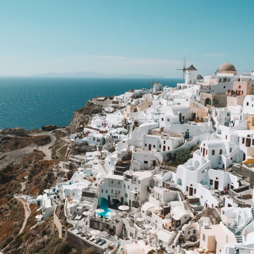 Cliffside village in Santorini with iconic whitewashed buildings, blue-domed roofs, a classic windmill, and a view of the calm Aegean Sea under a clear blue sky.