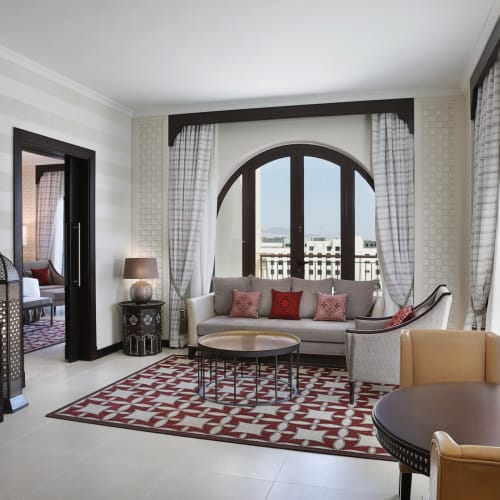 Elegant hotel living room with a white sofa, patterned pillows, and large arched windows revealing a view of buildings outside.