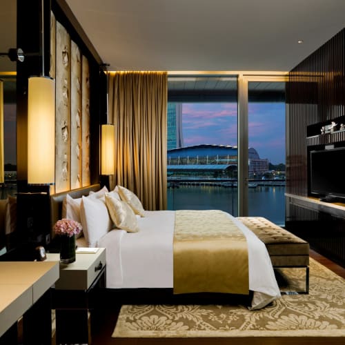Luxurious hotel suite with a large bed, view of a cityscape and water through floor-to-ceiling windows, elegant decor.