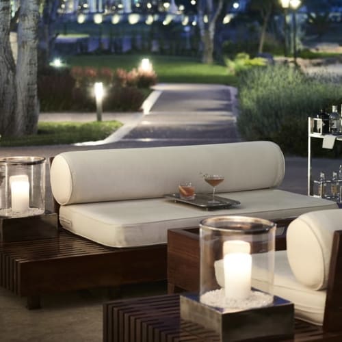 A luxurious outdoor lounge area with white cushioned seating, decorated with candles and surrounded by greenery at night.