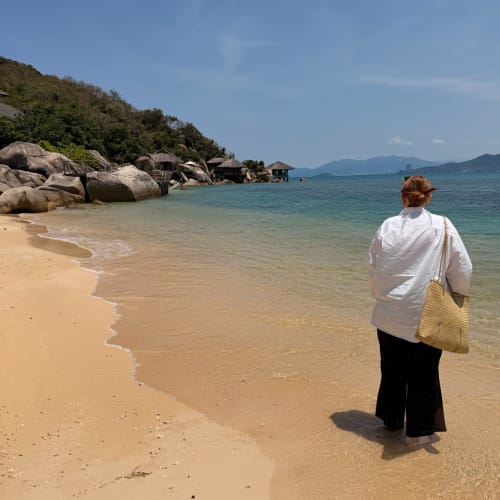 A person with short hair wearing a white shirt and black pants stands barefoot at the edge of clear seawater on a sandy beach, facing away, with rocky hillside and overwater bungalows in the distance under a blue sky.