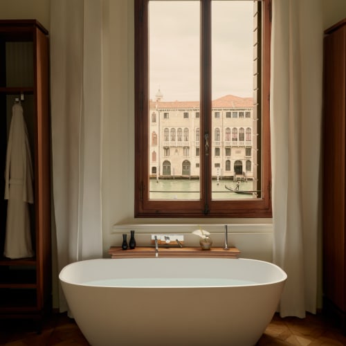 A luxurious bathroom with a white bathtub in front of a large window overlooking a canal and historic buildings.