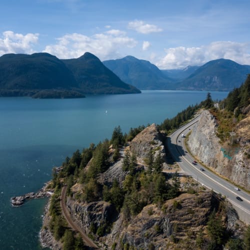 A scenic view of the Sea to Sky Highway winding along a mountain cliff beside a large lake, with forested slopes and distant mountain peaks under a partly cloudy sky.