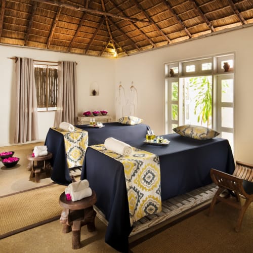 Ein ruhiger Spa-Raum mit drei Massagebetten, bedeckt mit dunkelblauen Laken, dekoriert mit gemusterten Handtüchern und umgeben von natürlichem Licht durch Fenster, mit Bambus- und Holz-Möbeln.
