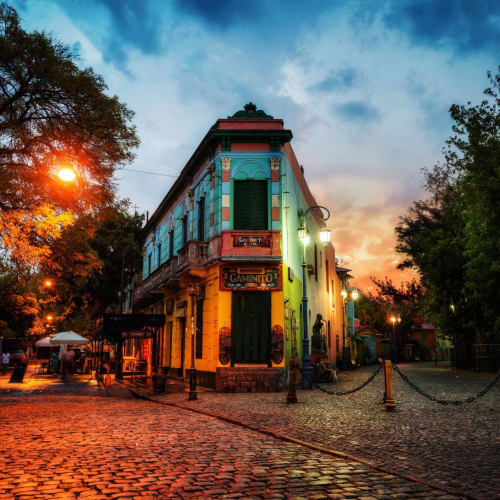 Bunte Gebäude säumen eine Pflasterstraße in La Boca, Buenos Aires bei Sonnenuntergang, mit Bäumen und Straßenlaternen.