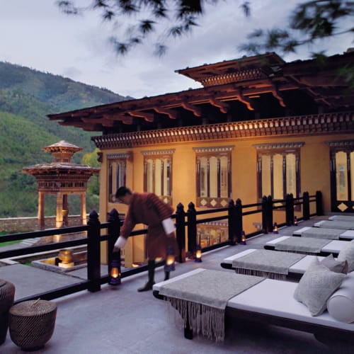 A person arranging cushions on a cozy outdoor terrace with traditional Bhutanese architecture and mountain scenery in the background.