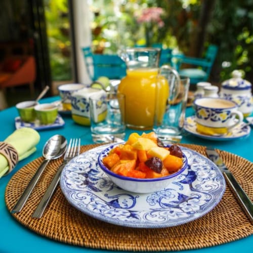 A colorful breakfast setup on a patio with a blue table, featuring a bowl of mixed fruit, a glass jug of orange juice, and various teacups and utensils.