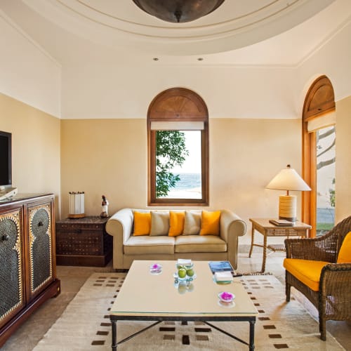 Elegant living room with a white sofa, wicker armchair, and a TV, with large windows showcasing a view of the sea.