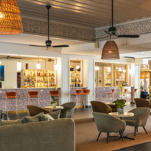 Innenraum einer stilvollen Bar mit einer langen Theke, die mit Ledersitzhockern bestückt ist, gemütlichen Sesseln um kleine runde Tische, hängenden geflochtenen Pendelleuchten und Deckenventilatoren in einer entspannten Lounge-Atmosphäre.