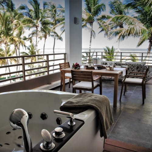 Außenbalkon mit einem Jacuzzi im Vordergrund und einem Esstisch mit Stühlen, umgeben von Palmen und Meerblick im Hintergrund.