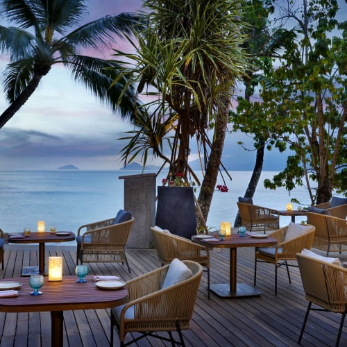Outdoor restaurant seating on a wooden deck overlooking the ocean, surrounded by palm trees and tropical plants at dusk with candles on the tables.
