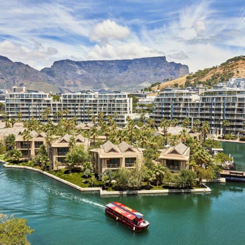 Luxurious waterfront resort with modern buildings and a boat sailing on a calm river surrounded by lush greenery in Cape Town, South Africa.