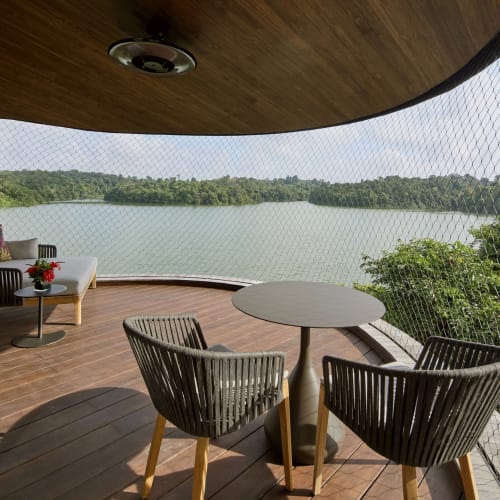 Eine Terrasse mit zwei Stühlen und einem runden Tisch mit Blick auf einen großen See, umgeben von üppigem Grün, eingezäunt mit einem Sicherheitsnetz unter einer Holzdecke.