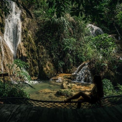 A person relaxing on a hammock in front of a lush waterfall in a jungle setting.