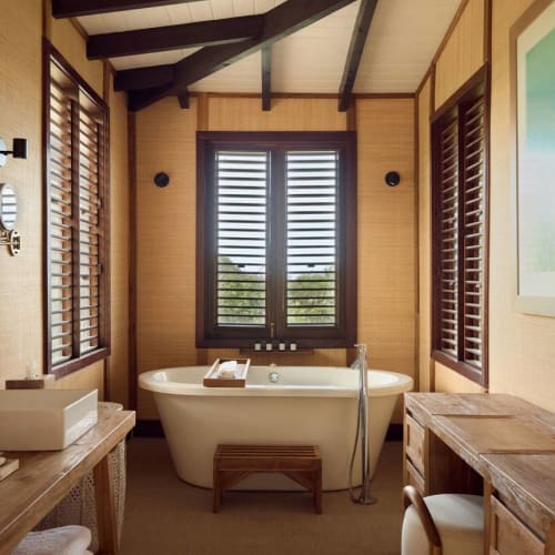 A cozy bathroom with a freestanding bathtub centered between two windows with wooden shutters, and a double sink with a wooden countertop.