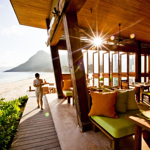 Eine Frau geht an einem Holzweg neben einem Strandrestaurant mit offenem Design entlang, mit Blick auf den Ozean und Berge im Hintergrund, Sonnenstrahlen fallen durch.