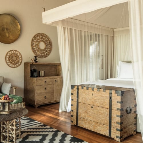 Ein ruhiges Schlafzimmer mit Himmelbett, das mit weißen, transparenten Vorhängen umhüllt ist, einer Holztruhe am Fußende des Bettes und einem Sitzbereich mit einem gemusterten Teppich, einem Sofa mit Kissen und dekorativen runden Wanddekorationen in einem tropischen Resort.