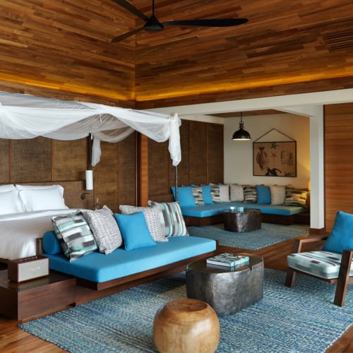 A spacious bedroom with a large bed draped in white fabric, surrounded by wooden walls and floors, featuring a seating area with blue and patterned cushions, and a small sitting nook with a chair and ottomans.