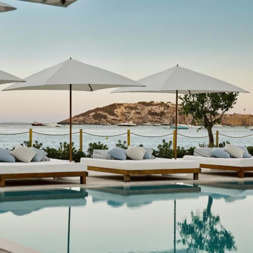 Poolside lounge chairs with large white umbrellas overlooking Ibiza Bay at sunset.