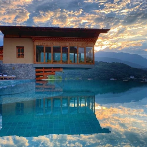 Eine luxuriöse Villa mit großen Glasfenstern spiegelt sich in einem Infinity-Pool, mit Bergen und einem dramatischen Sonnenuntergangshimmel in Bhutan.