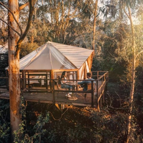 Two people sitting on a wooden deck outside a glamping tent surrounded by trees during sunset.
