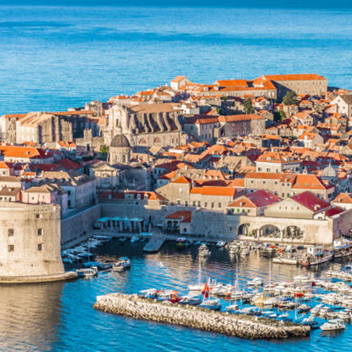 Eine Luftaufnahme von Dubrovnik mit historischen Mauern, terrakotta-gefliesten Gebäuden und einem Yachthafen an der Adriaküste.