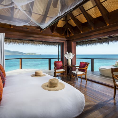 Luxurious bedroom with a large bed, surrounded by white curtains, overlooking the ocean from a wooden deck.