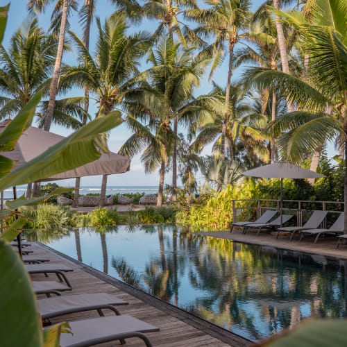 Ein Resort-Schwimmbecken, umgeben von Palmen und Liegestühlen, mit Sonnenschirmen und Meerblick im Hintergrund.