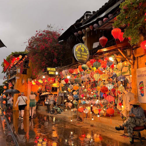 A rainy evening street scene in Hoi An, Vietnam, with colorful hanging lanterns illuminating a shop and pedestrians walking with umbrellas along the wet reflective pavement.
