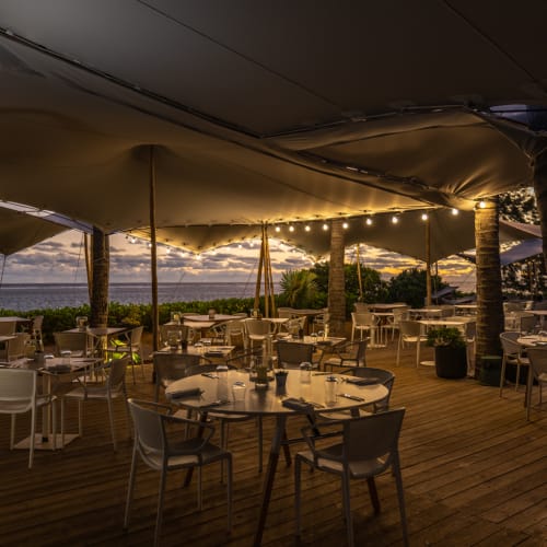 Ein Außenbereich eines Restaurants in der Dämmerung mit Lichterketten unter einem großen Sonnensegel, im Hintergrund der Ozean und Palmen.
