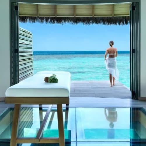 Eine Frau steht auf einer Holzveranda, blickt auf den Ozean, gesehen von innen eines luxuriösen Overwater-SPAs