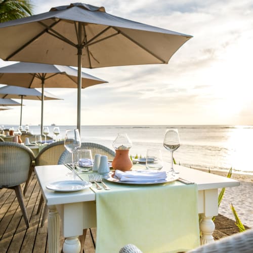 An elegantly set dining table with glassware and a table runner on a wooden deck by the beach, shaded by a large umbrella with ocean view at sunset.