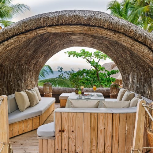 Ein gemütlicher privater Speiselounge-Bereich mit Holzbänken und Kissen unter einem gewölbten Strohdach, umgeben von tropischen Palmen und Grünpflanzen in einer ruhigen Außenumgebung.