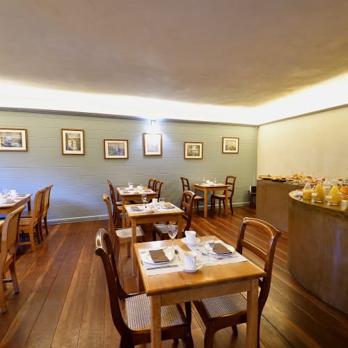 Ein kleiner, gemütlicher Frühstücksraum mit ordnungsgemäß arrangierten Holztischen und -stühlen, eingedeckt mit Tassen, Tellern und Besteck, und einem gebogenen Buffet-Tresen mit frischem Obst und Saftkannen auf der rechten Seite.