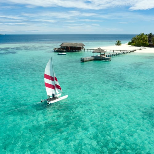 Ein Katamaran segelt über türkisfarbenes Wasser nahe einer kleinen Insel mit Überwasser-Bungalows auf Mirihi, Malediven.