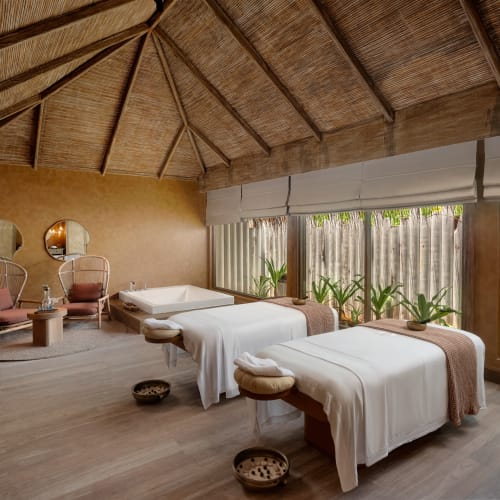 Ein ruhiger Spa-Raum mit zwei Massageliegen, bedeckt mit weißen Laken und braunen Decken unter einem Strohdach, mit großen Fenstern und Bambus-Jalousien sowie grünen Pflanzen außen, daneben zwei Stühle und ein kleiner Tisch.