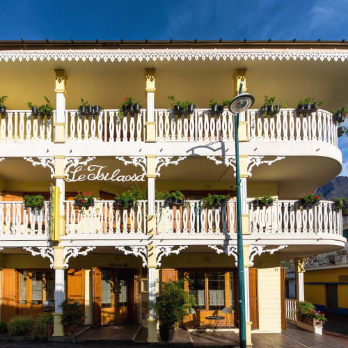 Dreistöckiges gelb-weißes Gebäude mit dekorativen Balkonen und Topfpflanzen auf jeder Ebene unter klarem blauem Himmel.
