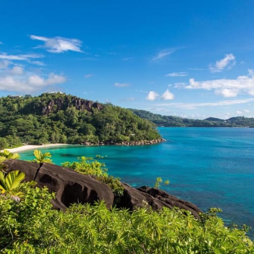 Eine malerische Aussicht auf eine tropische Küste mit üppigem Grün, felsigen Ufern und türkisem Wasser unter einem teils bewölkten Himmel.