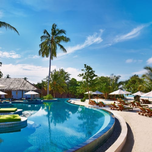 Ein luxuriöser Hotelpool, umgeben von Palmen und Cabanas an einem tropischen Strand mit klarem blauem Wasser und Himmel.