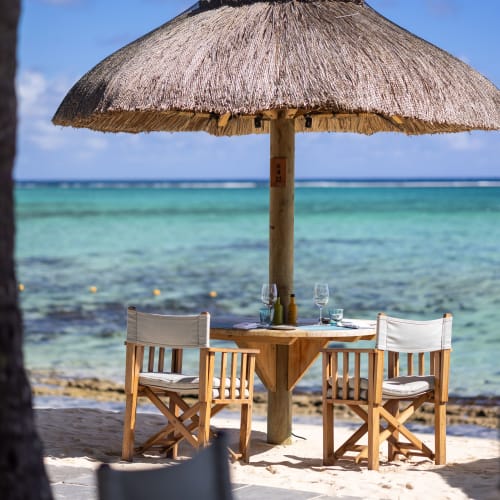 A tropical beach setting featuring a wooden table with two cushioned chairs under a large thatched umbrella, overlooking clear turquoise waters.