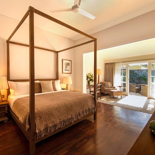 A bedroom with a four-poster bed, two bedside lamps, and framed artwork on the wall, leading to a sitting area with chairs and large windows with curtains.