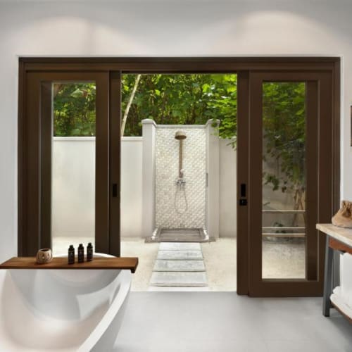 A luxurious bathroom with a large bathtub and an outdoor shower area separated by sliding glass doors, overlooking a garden.