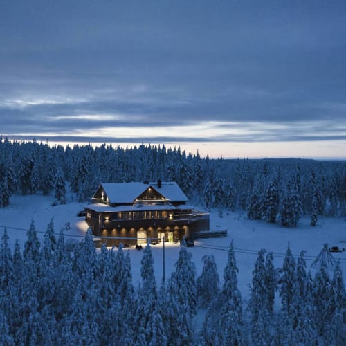 Eine verschneite Winterabend-Szene mit einer warm beleuchteten Lodge, umgeben von dicht mit Schnee bedeckten Kiefern unter einem bewölkten blauen Himmel.