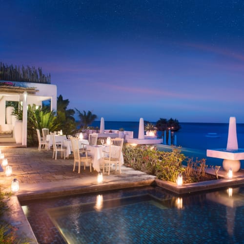 Elegantes Outdoor-Restaurant mit weißen Tischen und Stühlen neben einem Pool mit Blick auf den Ozean bei Dämmerung.