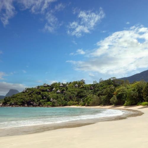 Ein unberührter Strand mit weißem Sand, türkisfarbenem Wasser, üppigem Grünland und Hügeln im Hintergrund unter einem teilweise bewölkten Himmel.