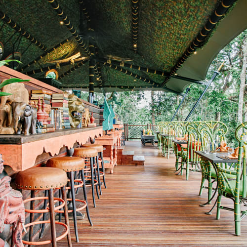 Eine hölzerne Außenterrasse eines Restaurants, umgeben von üppigem Grün, dekoriert mit traditionellen Artefakten, Statuen und lebendigen grünen Stühlen und Tischen.